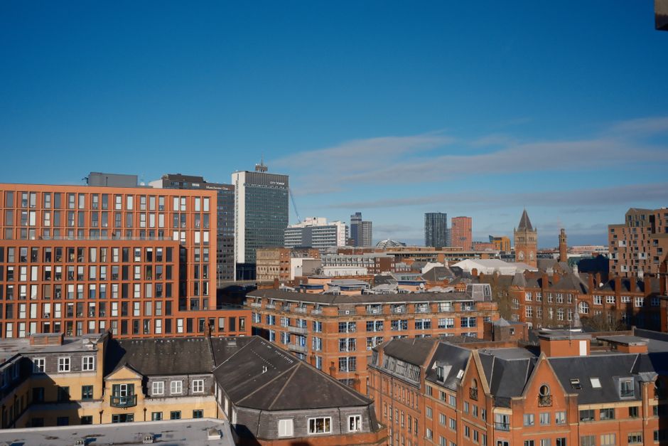 A cityscape featuring a mix of modern and historic buildings under a clear blue sky. In the foreground, there are residential flats with sloped grey roofs and brick facades, some with small windows. B